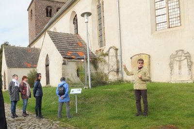 Man vor einer Kirche erläutert, daneben steht eine Infotafel.