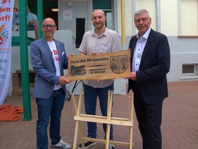 Drei Männer halten eine Holztafel mit der Aufschrift "Haus des Ehrenamtes" in der Hand.